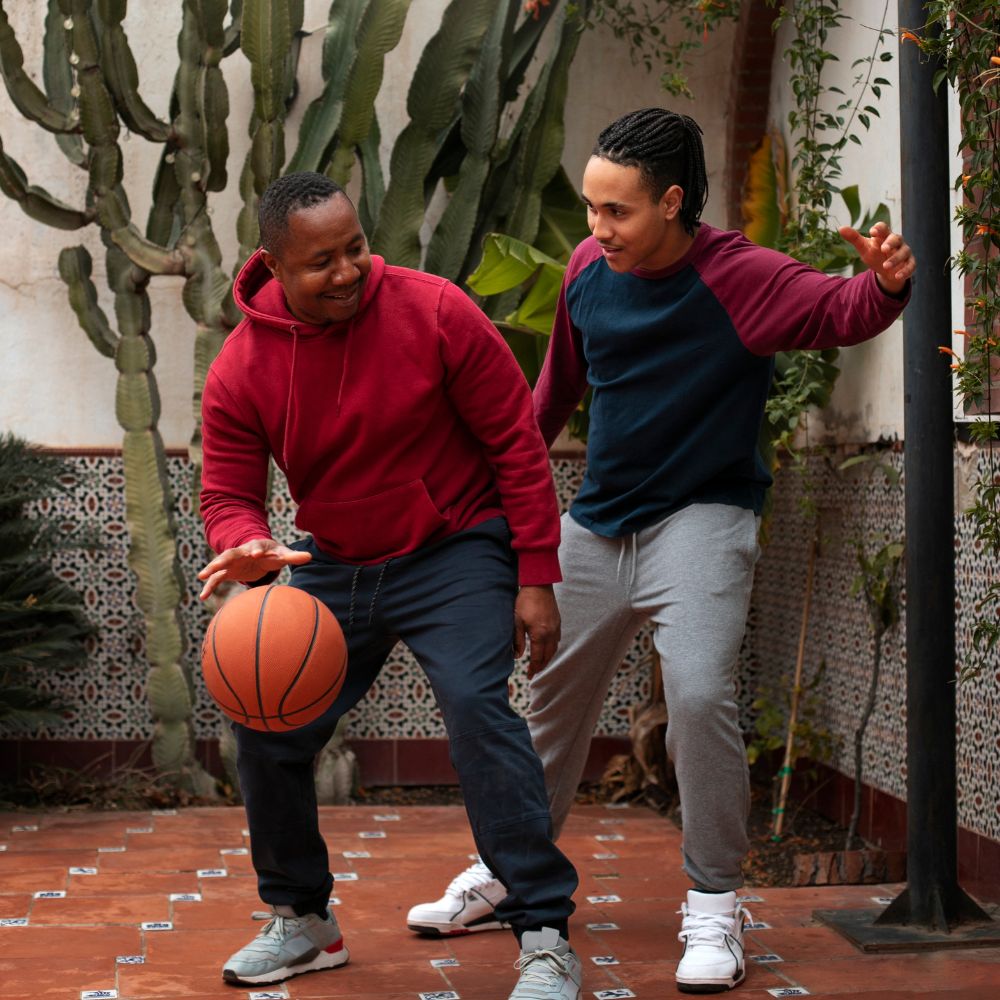 Two men playing with a basketball in an outdoor setting with plants and a wall.

