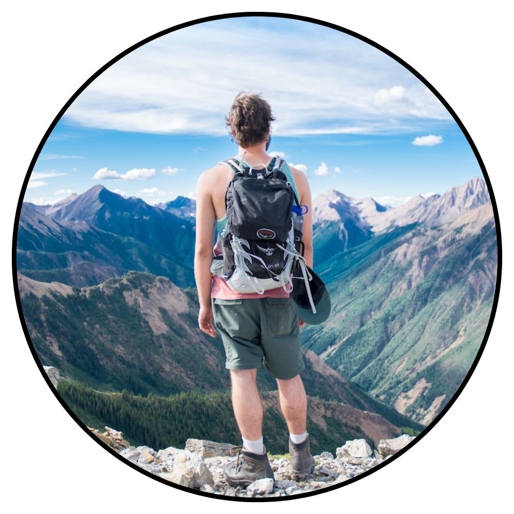 Person with a backpack standing on a mountain peak looking at a scenic view of mountains.

